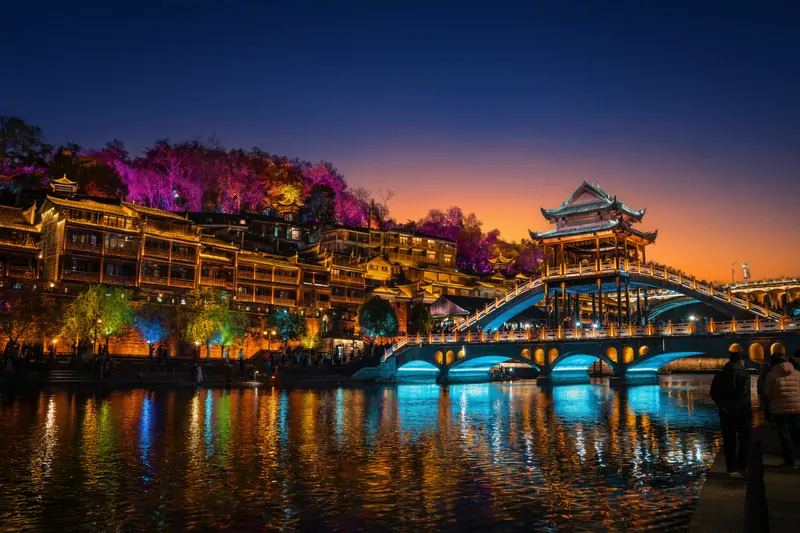 Древний город Фэнхуан (Fenghuang Ancient Town) в Китае, известный своими живописными видами. Фотография запечатлела ночную иллюминацию города, когда огни зданий отражаются в водах реки Туоцзян.